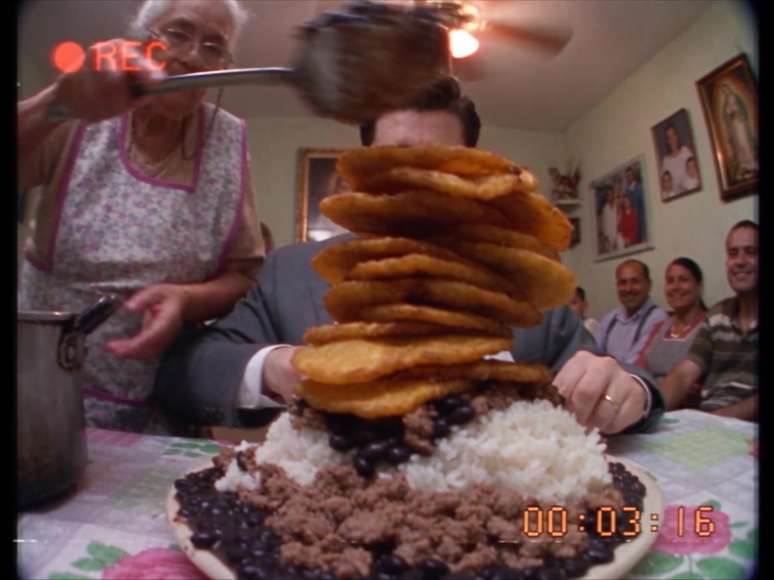 VHS still: a grandmother piles a tower of tortillas onto the plate of a man in a grey suit while the family watches.