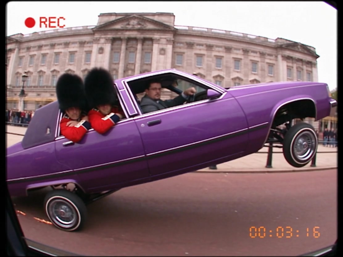 VHS still: a purple lowrider outside Buckingham Palace, the man in a grey suit at the wheel.