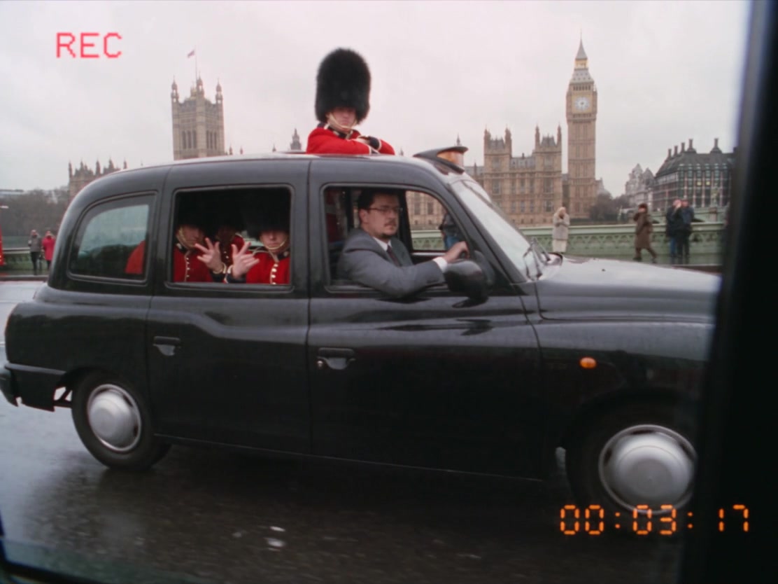VHS still: a black London taxi packed with Royal Guards in front of Big Ben, the man in a grey suit at the wheel.